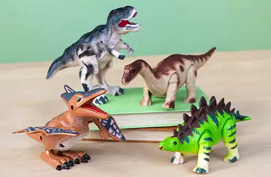 A collection of four toy dinosaurs is displayed on a wooden surface in front of a green background. The toys include a green and brown Stegosaurus, an orange Pteranodon with patterned wings, a brown and beige Brachiosaurus, and a grey and white Tyrannosaurus rex with its mouth open.