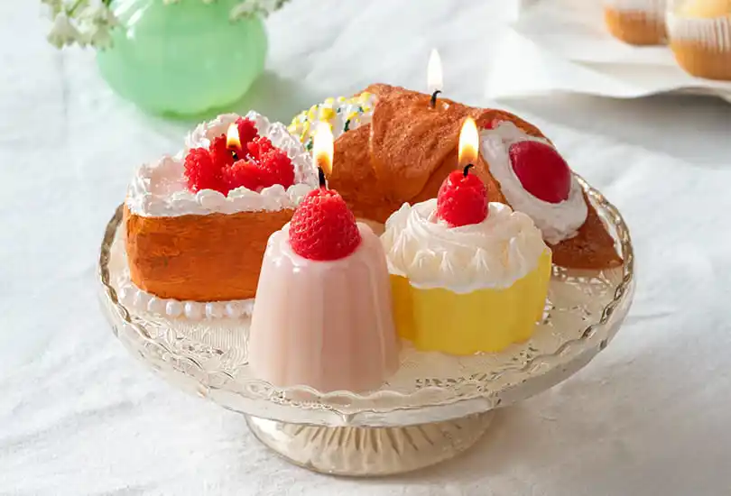 A plate of realistic dessert-shaped candles displayed on a glass cake stand, including cakes and puddings topped with strawberries and cream-like decorations.