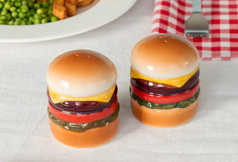 Two ceramic salt and pepper shakers shaped like hamburgers displayed on a table next to a plate of food and a red‑and‑white checked napkin.