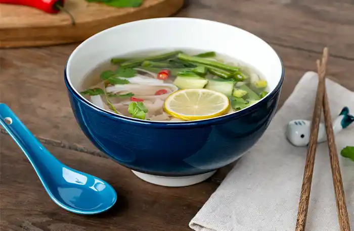 Ceramic noodle bowl with a dark blue and white glaze, paired with a light blue ceramic spoon, wooden chopsticks, and a small whale‑shaped ceramic chopstick rest.