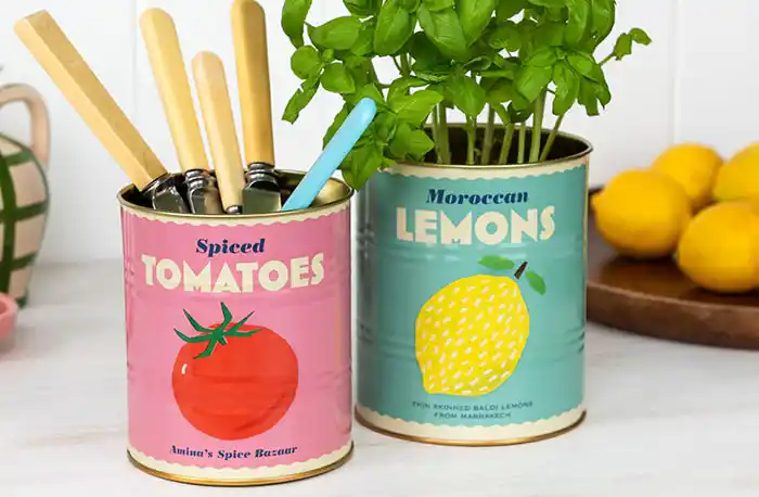 Two colorful tin cans repurposed as containers on a white surface. The left can is pink with the text “Spiced Tomatoes” and an illustration of a red tomato, holding kitchen utensils. The right can is teal with the text “Moroccan Lemons” and an illustration of a yellow lemon, containing green leafy herbs. A lemon is partially visible in the background.