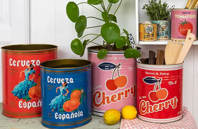 Collection of large decorative tins with colourful vintage-style designs, including red and blue tins labelled ‘Cerveza Española’ with illustrations of oranges and a dancer, and pink and red tins labelled ‘Cherry Sweet & Juicy.’ The tins are used as storage for plants and kitchen utensils, with lemons placed in front and smaller tins of olive oil and tomatoes on a shelf in the background.
