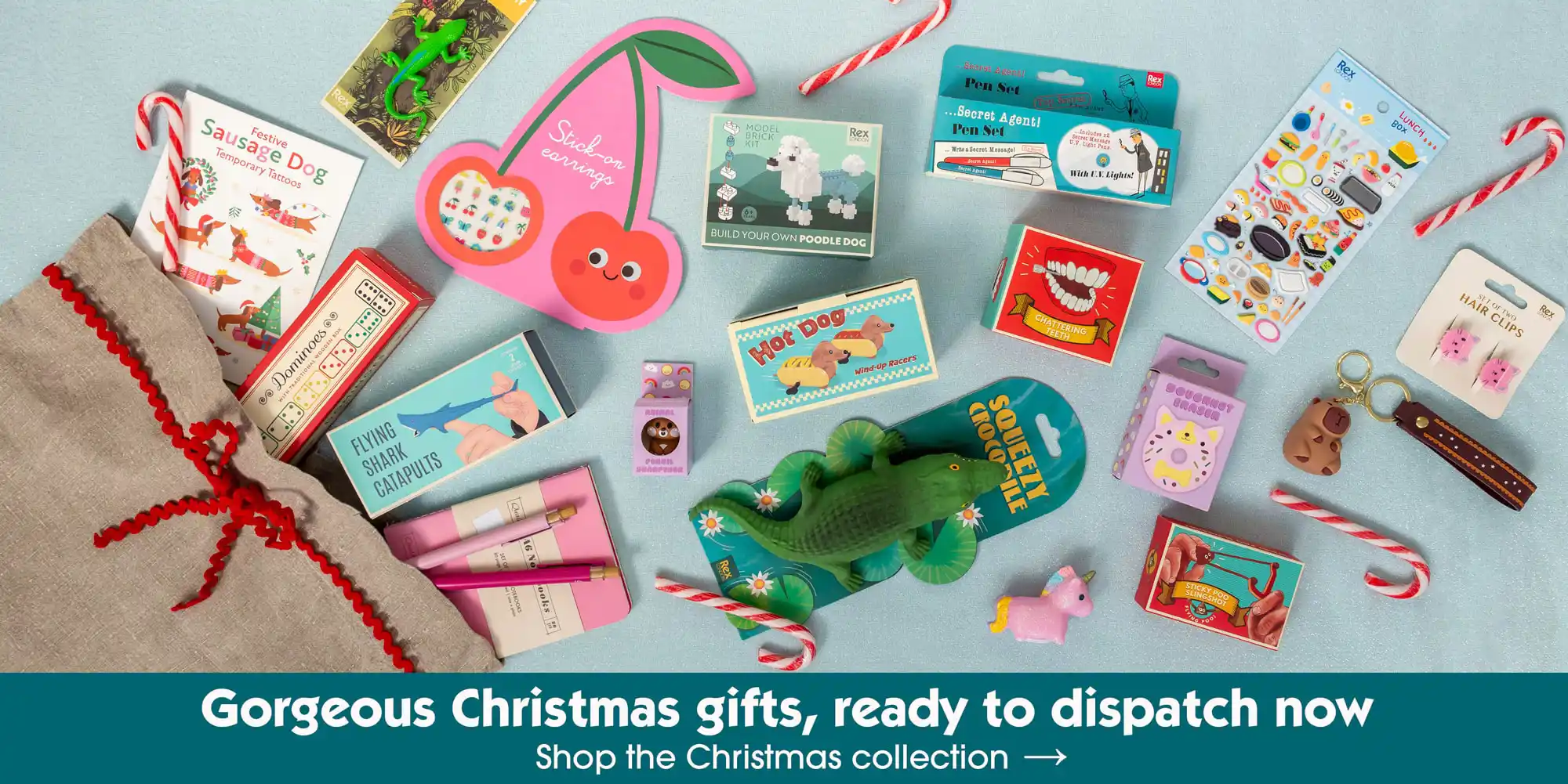 Flat lay of assorted stocking filler gifts on a light blue background, including candy canes, novelty toys, stickers, mini games, and beauty items.