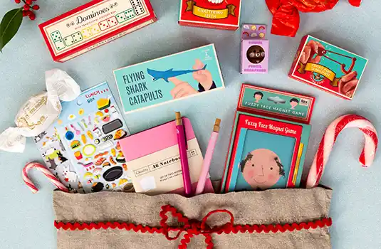 Assortment of small gift items including mini games, pencils, candy canes, and colourful packaging, displayed with a linen bag tied with red ribbon