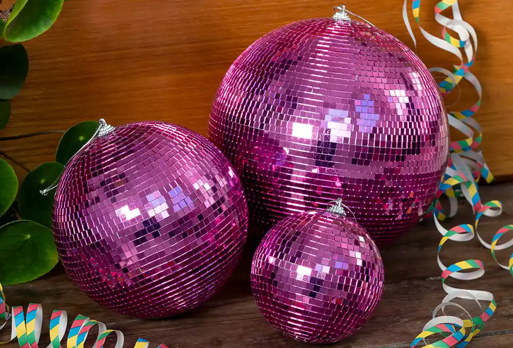 Three shiny pink disco balls of different sizes placed on a wooden surface. The disco balls have a reflective mosaic pattern that creates a sparkling effect.