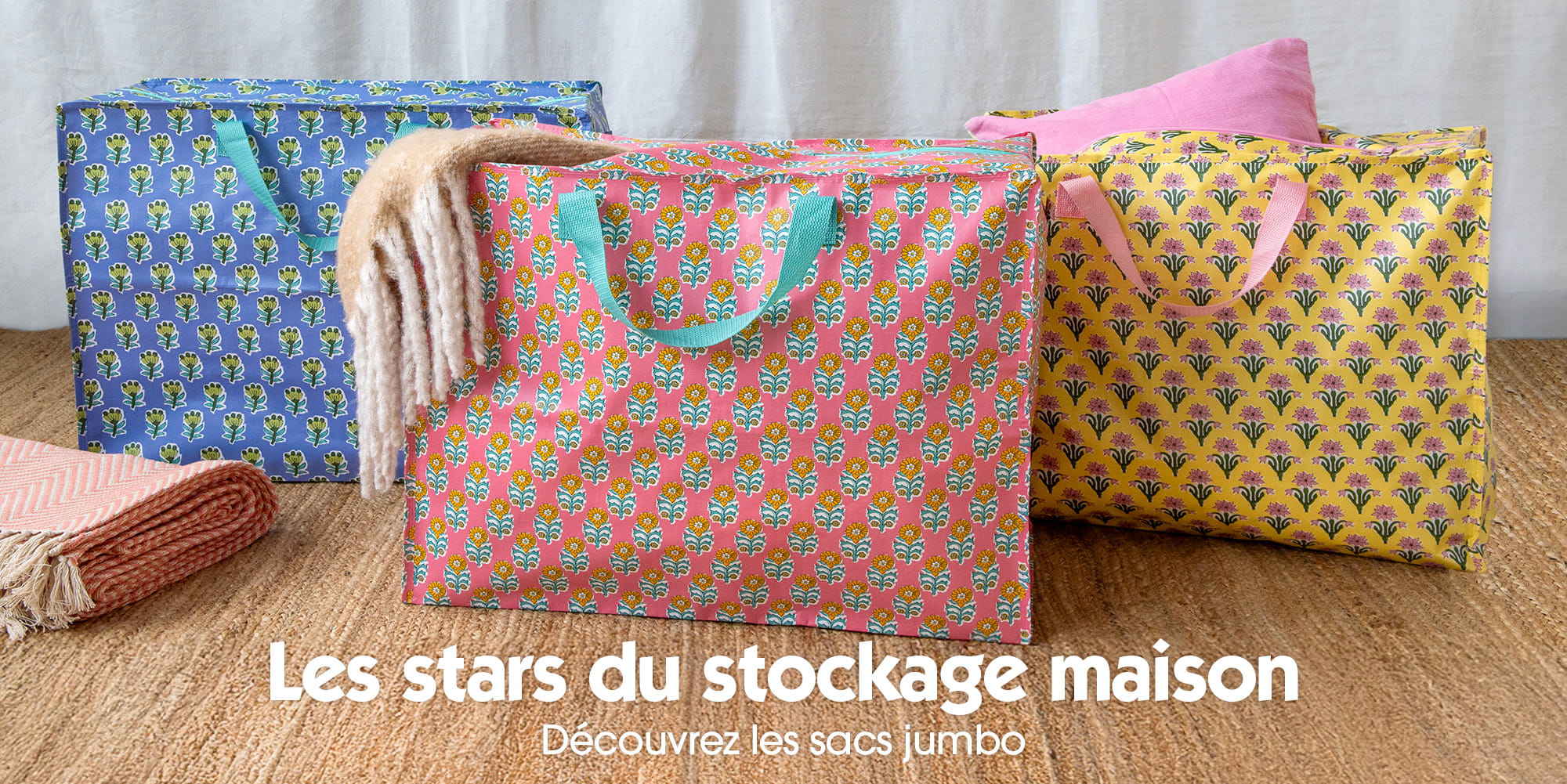 Three large patterned storage bags in blue, pink and yellow displayed on a woven mat, with folded blankets placed beside them.