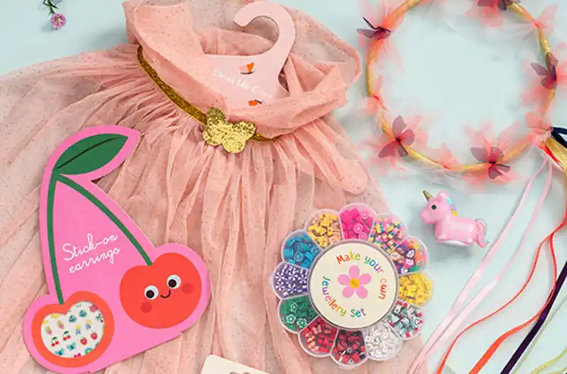 Children’s accessories including a pink dress, headband with ribbons, unicorn toy, and beaded jewellery set arranged on a pastel background.