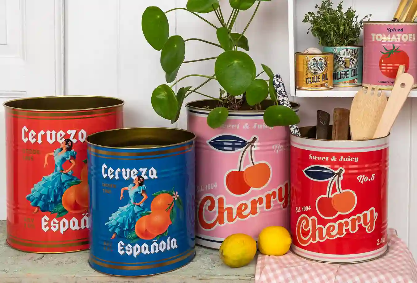 Collection of large decorative tins with colorful vintage-style designs, including red and blue tins labeled ‘Cerveza Española’ with illustrations of oranges and a dancer, and pink and red tins labeled ‘Cherry Sweet & Juicy.’ The tins are used as storage for plants and kitchen utensils, with lemons placed in front and smaller tins of olive oil and tomatoes on a shelf in the background.