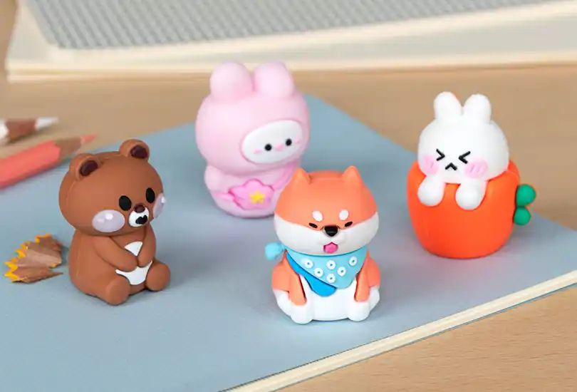 Four small, cute animal figurines in bright colours, including a bear, bunny, fox, and white bunny in an orange cup, displayed on a pastel blue notebook.