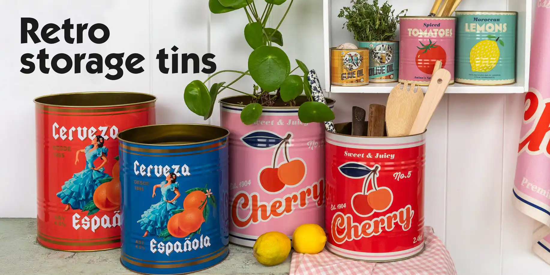 Collection of colorful decorative tins with vintage-style designs, including red and blue tins labeled ‘Cerveza Española’ and pink and red tins labeled ‘Cherry.’ The tins are arranged on a countertop with lemons, wooden utensils, and a plant. Additional smaller tins labelled ‘Tomatoes’ and ‘Lemons’ are displayed on a shelf.
