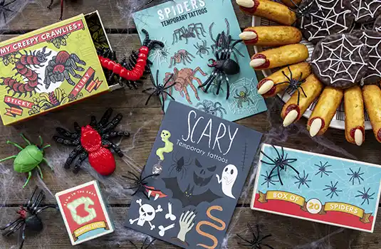 Halloween-themed flat lay featuring spider and creepy crawly toys, boxes of temporary tattoos, plastic insects, and a spider web-decorated dessert tray with Twinkie-style treats.