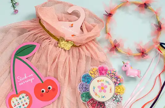 Children’s accessories including a pink dress, headband with ribbons, unicorn toy, and beaded jewellery set arranged on a pastel background.