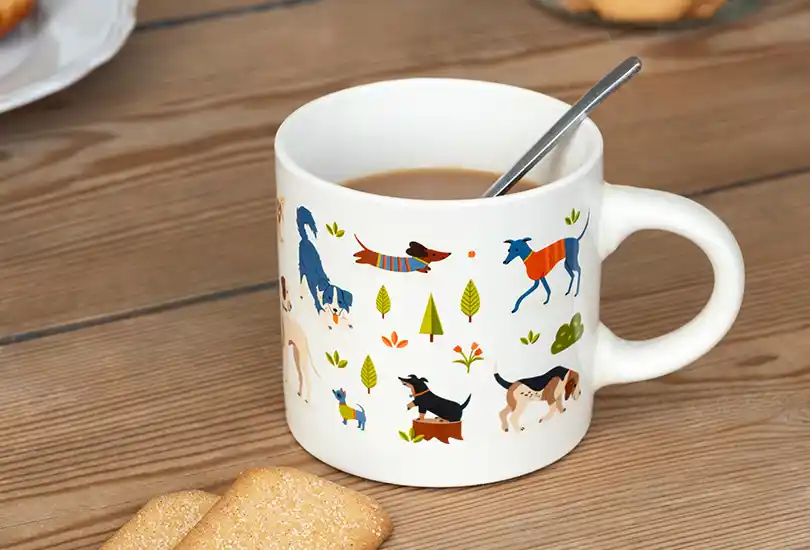 A white ceramic mug. The mug is decorated with colourful illustrations of various dog breeds in different poses, along with small green trees and orange floral accents.