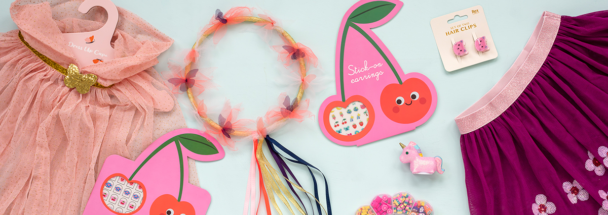 Children’s dress-up accessories on a light blue background, including a pink dress with a gold belt and butterfly detail, a butterfly headband, stick-on earrings, pink cat hair clips, a purple floral skirt, a unicorn figurine, and colourful hair ties.