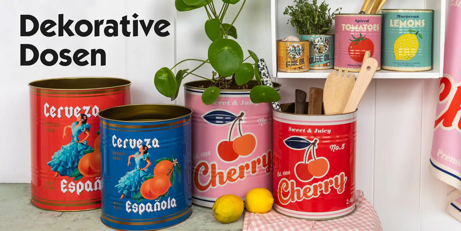 Collection of colorful decorative tins with vintage-style designs, including red and blue tins labeled ‘Cerveza Española’ and pink and red tins labeled ‘Cherry.’ The tins are arranged on a countertop with lemons, wooden utensils, and a plant. Additional smaller tins labelled ‘Tomatoes’ and ‘Lemons’ are displayed on a shelf.