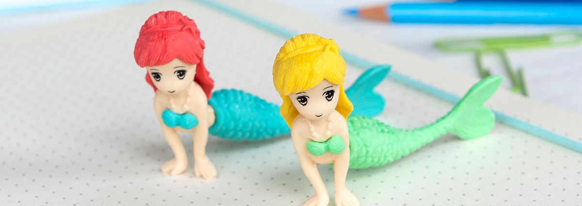Two small mermaid figurines with turquoise tails on a dotted notebook, one with red hair and one with yellow hair, alongside a blue pencil and green paper clips.