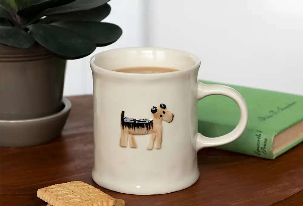 Cream-coloured ceramic mug filled with tea, featuring a raised design of a small dog on the front. 