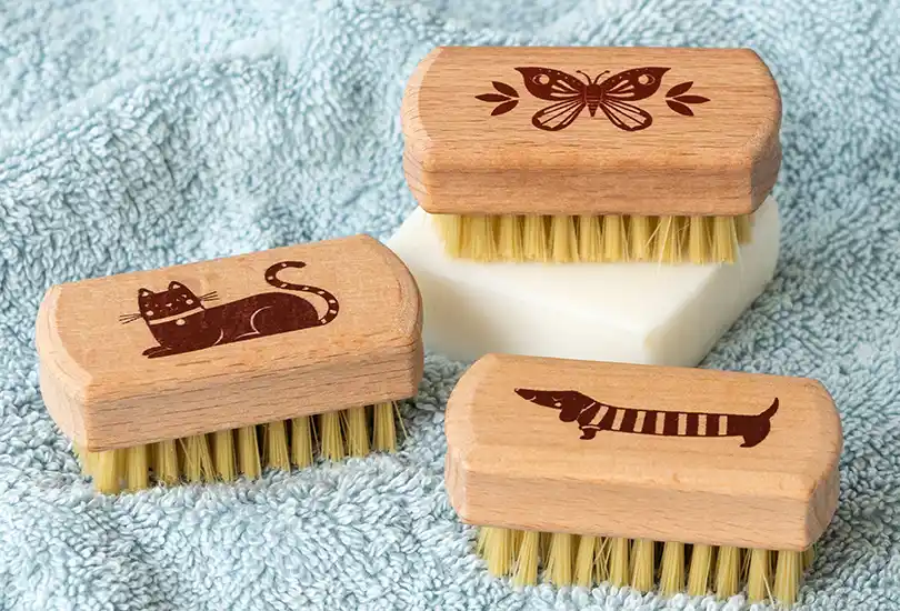Three mini wooden nail brushes with engraved designs of a cat, a butterfly, and a sausage dog.