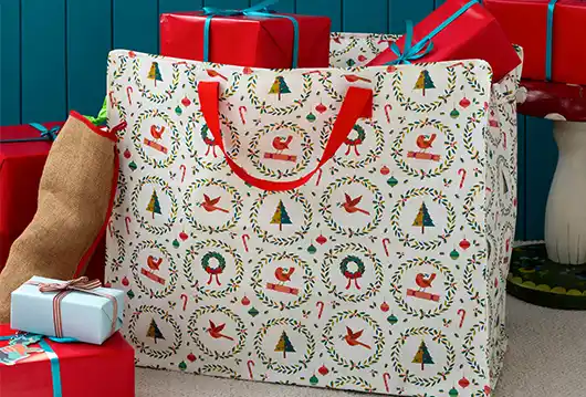 Large festive storage bag with red handles and Christmas-themed print featuring Santa, wreaths, trees, and candy canes, surrounded by wrapped gifts in red and blue paper with ribbons.