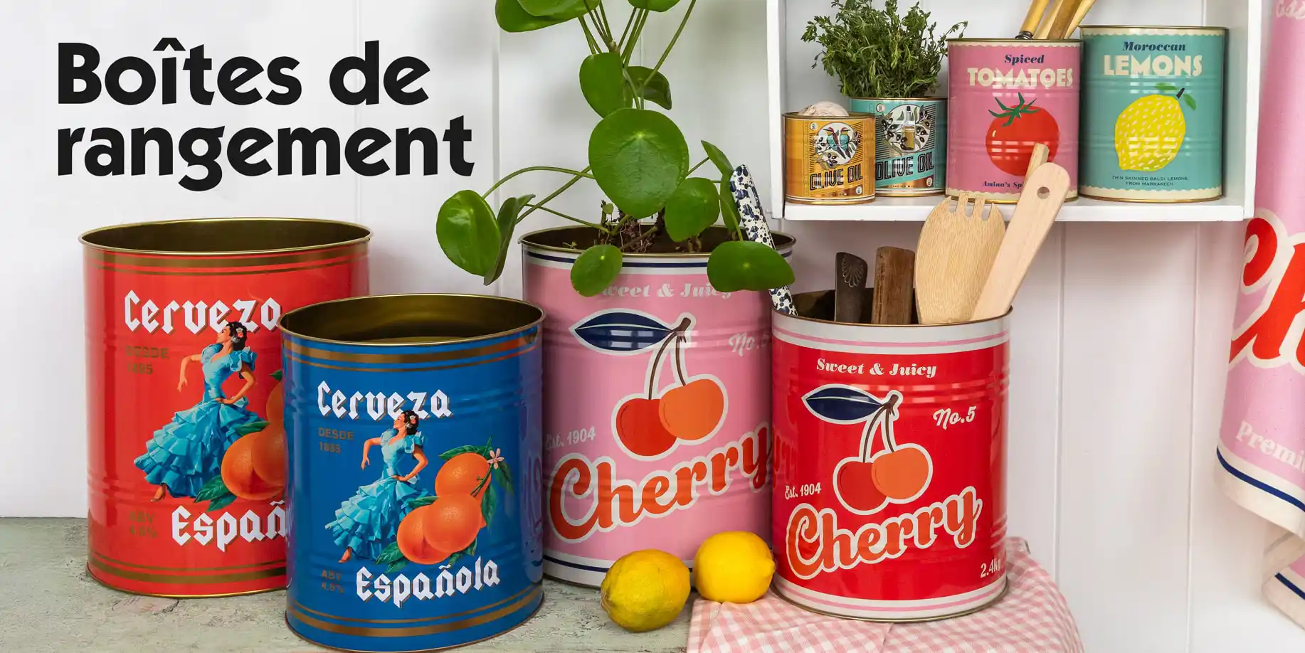 Collection of colorful decorative tins with vintage-style designs, including red and blue tins labeled ‘Cerveza Española’ and pink and red tins labeled ‘Cherry.’ The tins are arranged on a countertop with lemons, wooden utensils, and a plant. Additional smaller tins labelled ‘Tomatoes’ and ‘Lemons’ are displayed on a shelf.