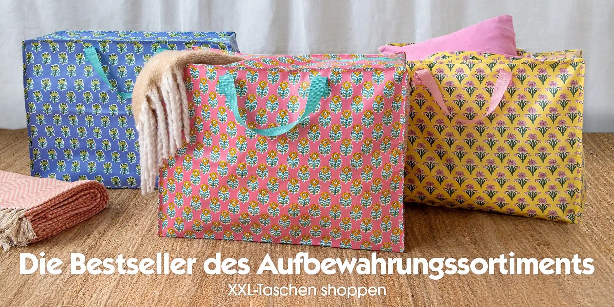 Three large patterned storage bags in blue, pink and yellow displayed on a woven mat, with folded blankets placed beside them.