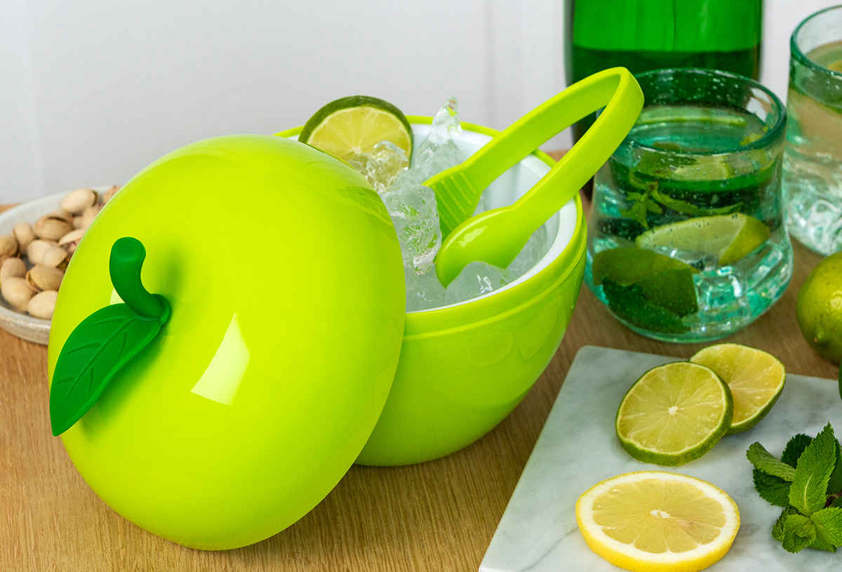 Green apple-shaped ice bucket with tongs, ice, and lime, on a table with citrus slices, mint, and drinks.