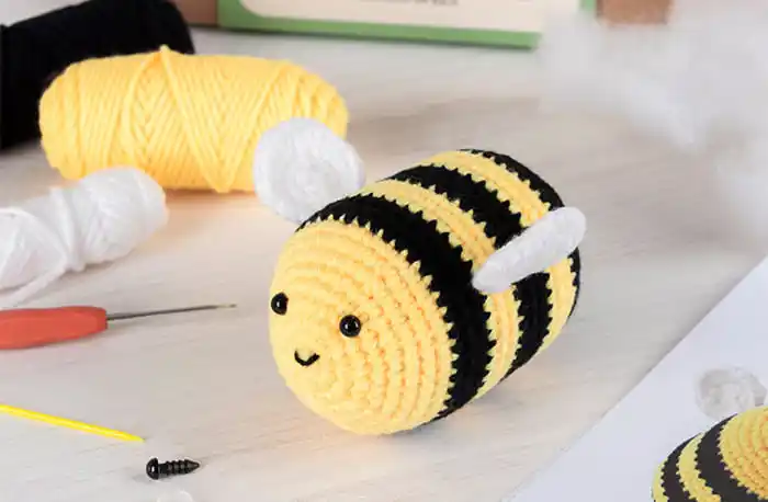 A crocheted bee plush toy with black and yellow stripes, white wings, and a smiling face.