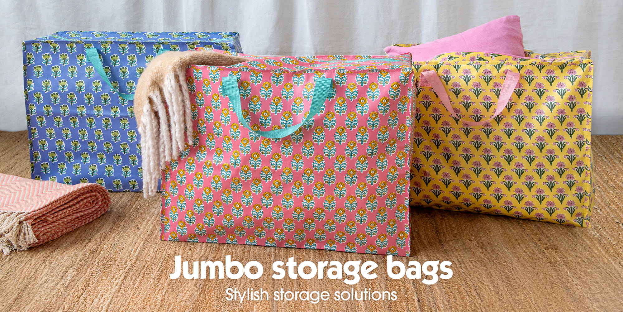 Three large patterned storage bags in blue, pink and yellow displayed on a woven mat, with folded blankets placed beside them.