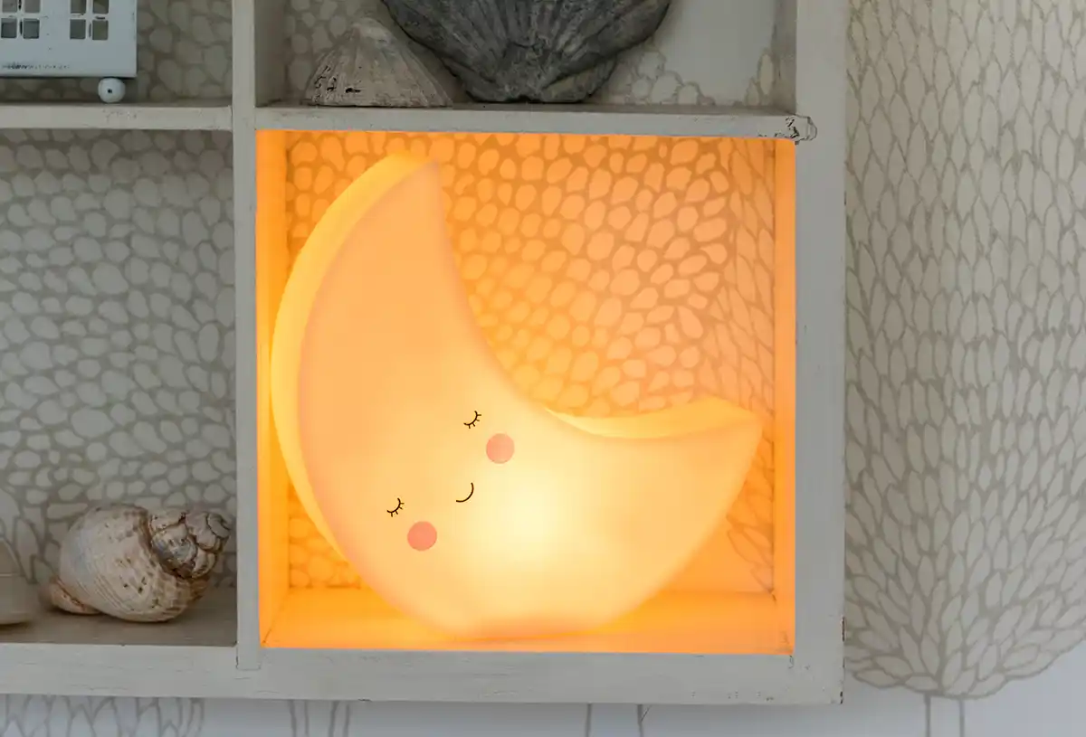 A glowing crescent moon-shaped lamp with a smiling face and rosy cheeks, placed inside a white shelf with seashell decorations.