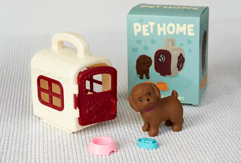 A cream-colored pet carrier with a red door and windows, and a small brown toy puppy wearing a purple collar. also in front of the puppy are two mini food bowls,  one pink and one blue with dog bone.